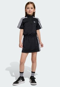Čierne športové šaty s krátkymi rukávmi, zipsovým golierom, elastickým pásom, bielymi bočnými pásmi, v kombinácii s čiernymi teniskami a pruhovanými ponožkami.