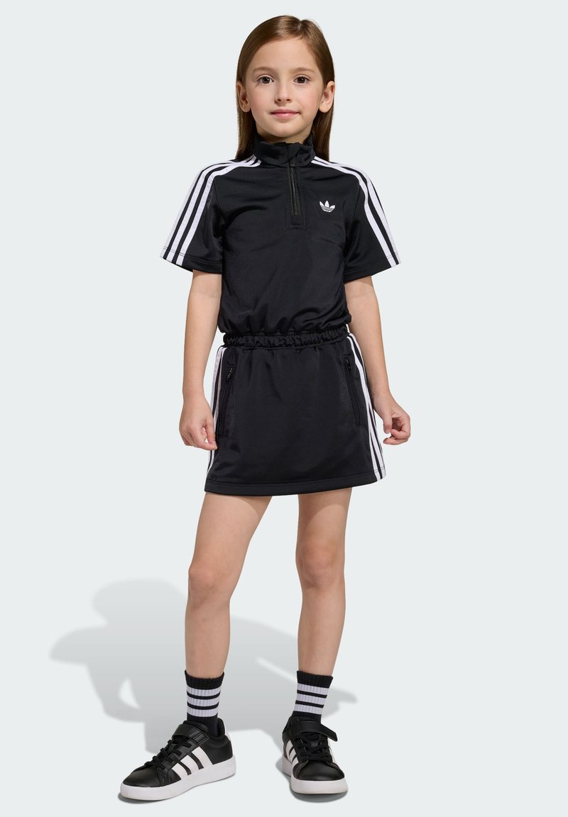 Čierne športové šaty s krátkymi rukávmi, zipsovým golierom, elastickým pásom, bielymi bočnými pásmi, v kombinácii s čiernymi teniskami a pruhovanými ponožkami.