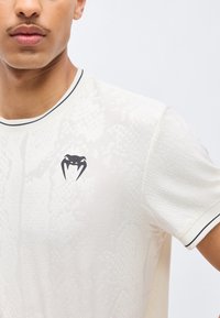 Νέος άνδρας που φοράει λευκό μπλουζάκι με υφή, με μαύρο λογότυπο κεφαλής φιδιού στο αριστερό στήθος και μαύρο περίγραμμα στο γιακά και τα μανίκια.