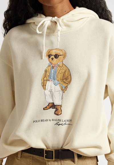 Sweatshirt creme com capuz com estampa de um urso usando óculos de sol, casaco castanho, camisola azul, calças brancas e botas castanhas; inclui texto com o nome da marca abaixo.
