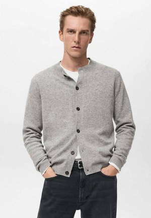 Cárdigan de punto gris con cuello redondo, botones frontales y puños de canalé. Llevado sobre una camisa blanca y combinado con jeans negros.