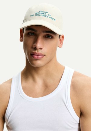 Cream baseball cap with green embroidered text reading "PROUD MEMBER OF THE EXCLUSIVE CREW." Worn with a fitted white tank top.
