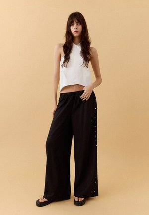 Joven mujer con cabello largo y castaño lleva un top corto blanco sin mangas, pantalones anchos negros con botones laterales y sandalias negras con plataforma, de pie.