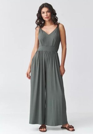 Kobieta stojąca w bezrękawnym szarym kombinezonie z szerokimi nogawkami i brązowych sandałach na jasnym, jednolitym tle.