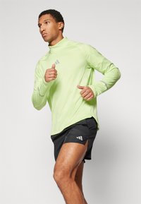 Hellgrünes, langärmliges Sportoberteil aus strukturiertem Stoff mit grauen Akzenten, kombiniert mit schwarzen Shorts mit seitlichem Schlitz und Logodetails.
