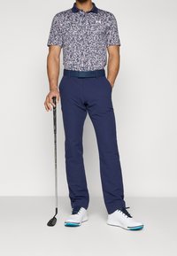 Marinblå blommig polo med korta ärmar, matchad med marinblå byxor. Vita golfskor med blå detaljer. Golfklubba i handen.