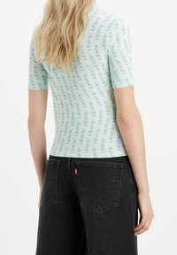 Světle modrý top s krátkým rukávem, na kterém je opakující se logo "LEVI'S" uspořádané ve vertikálním vzoru. V kombinaci s černými džíny s ikonickým designem zadní kapsy.