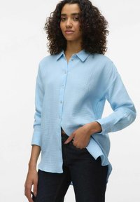 Light blue button-up shirt in textured fabric, featuring a collar, long sleeves, and a relaxed fit. Paired with dark denim jeans.