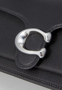 Borsa a tracolla in pelle nera con un grande fermaglio in metallo argentato, cuciture bianche e una superficie texture.