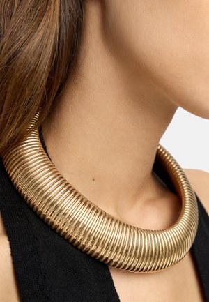 Gruby, żebrowany złoty choker noszony na szyi kobiety o prostych brązowych włosach i czarnym, bezrękawnym topie.