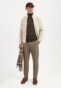 Beige fleece jacket, donkerbruine coltrui, lichtbruine broek, bruin schoenen en een geruit sjaal, staand tegen een eenvoudige witte achtergrond.