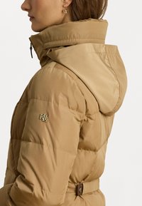 Tömött kabát homokszínben, magas gallérral és kapucnival. Tűzött mintázattal rendelkezik, és arany logó díszítéssel a ujján. Texturált, sima anyag.