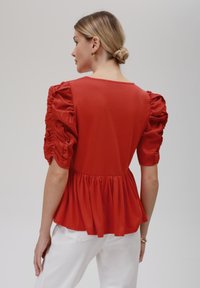 Rød peplum-top med puffede ærmer, lavet af blødt stof. Har en rund halsudskæring og samlede detaljer i taljen.