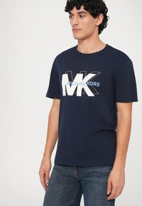 Hombre vistiendo una camiseta navy blue de Michael Kors con un gran logo "MK" en blanco y naranja, y jeans azules deslavados, de pie contra un fondo blanco.