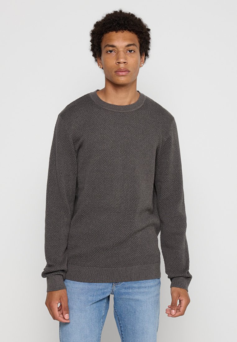 Suéter de punto texturizado gris con cuello redondo y puños acanalados, combinado con jeans azul claro, exhibido sobre un fondo neutral.