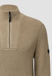 Beżowy sweter z dzianiny z wysokim kołnierzem i zamkiem błyskawicznym z przodu, z teksturowanymi wzorami ściągaczy i wafli oraz małą czarną łatką na rękawie.