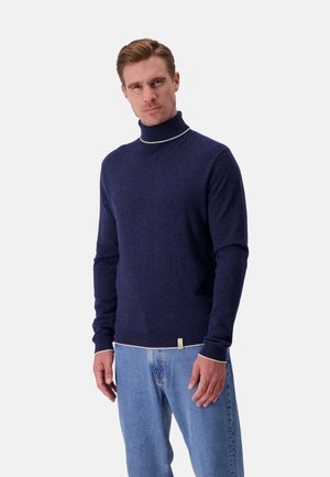 Marineblauer Rollkragenpullover mit weißem Akzent am Kragen, aus einer Wollmischung, mit langen Ärmeln und taillierter Passform. Kombiniert mit blauen Jeans.