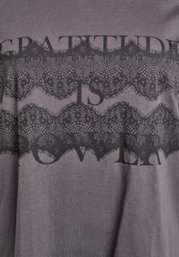 Graues Baumwoll-T-Shirt mit einem schwarzen Spitzenmuster und dem Text "DANKBARKEIT IST KRAFT" in fetten Buchstaben, was einen kontrastierenden Effekt erzeugt.