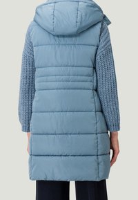 Padded vest in light blue with a hood, featuring a quilted design and textured knitted sleeves in a matching shade.