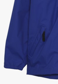 Giacca impermeabile blu con una texture liscia, dotata di una tasca laterale angolata e polsini elasticizzati. Ideale per condizioni di bagnato.