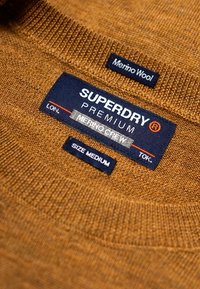 Pull en laine mérinos marron à col rond avec encolure côtel ée. Les étiquettes indiquent « Superdry Premium », « Merino Crew » et « Taille M ».
