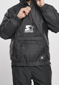 Black anorak jacket made of lightweight, shiny material. Features a quarter zip, elastic cuffs, front pocket, and white logo on chest.