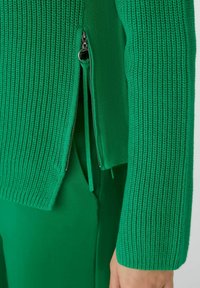 Pull en tricot vert avec un design texturé à côtes, doté d'une fermeture éclair sur le côté et d’une coupe décontractée. Les accents comprennent une doublure intérieure lisse.