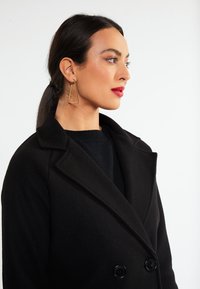 Lang sort uldfrakke med dobbelte knapper, lagt krave, parret med geometriske guldøreringe, sat mod en simpel hvid baggrund.