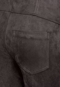 Close-up van donkerbruine suède broek waarin de achterzak en de gedetailleerde stiksels op de stof zichtbaar zijn.