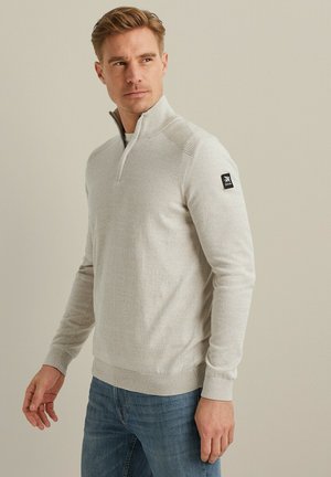 Lichtgrijze, gebreide pullover met een opstaande kraag, halve rits en subtiele ribgebreide accenten op de schouders; gedragen over een blauwe spijkerbroek.