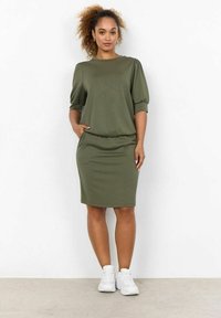 Vestido verde oliva con escote redondo, mangas cortas abullonadas, forma ajustada y bolsillos laterales, combinado con zapatillas blancas. Textura de tela suave.