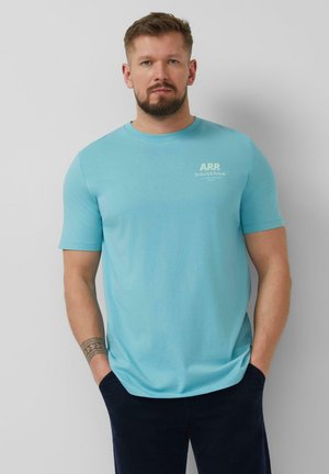 Homme avec une barbe portant un t-shirt bleu clair avec le logo A.R.R. et un pantalon foncé, debout devant un fond gris clair uni. Mains dans les poches.