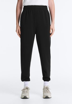 Persoană purtând pantaloni negri cu talie elastică și tiv la glezne, șosete albe, adidași bej și o cămașă albă pe un fundal alb simplu.