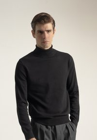 Hombre con el cabello peinado hacia atrás, vistiendo un suéter negro de cuello alto y pantalones grises con pliegues, de pie frente a un fondo claro y liso.