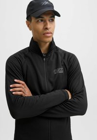 Chaqueta negra con cremallera y tejido texturizado, con cuello alto; presenta el logo "CNZ" en el pecho izquierdo; se lleva con los brazos cruzados.