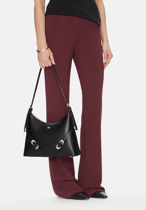 Mujer con pantalones burdeos y zapatos negros, sosteniendo un bolso de cuero negro con herrajes dorados por la correa.