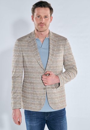 Man wearing beige plaid blazer over light blue polo shirt and blue jeans, buttoning the blazer with one hand.