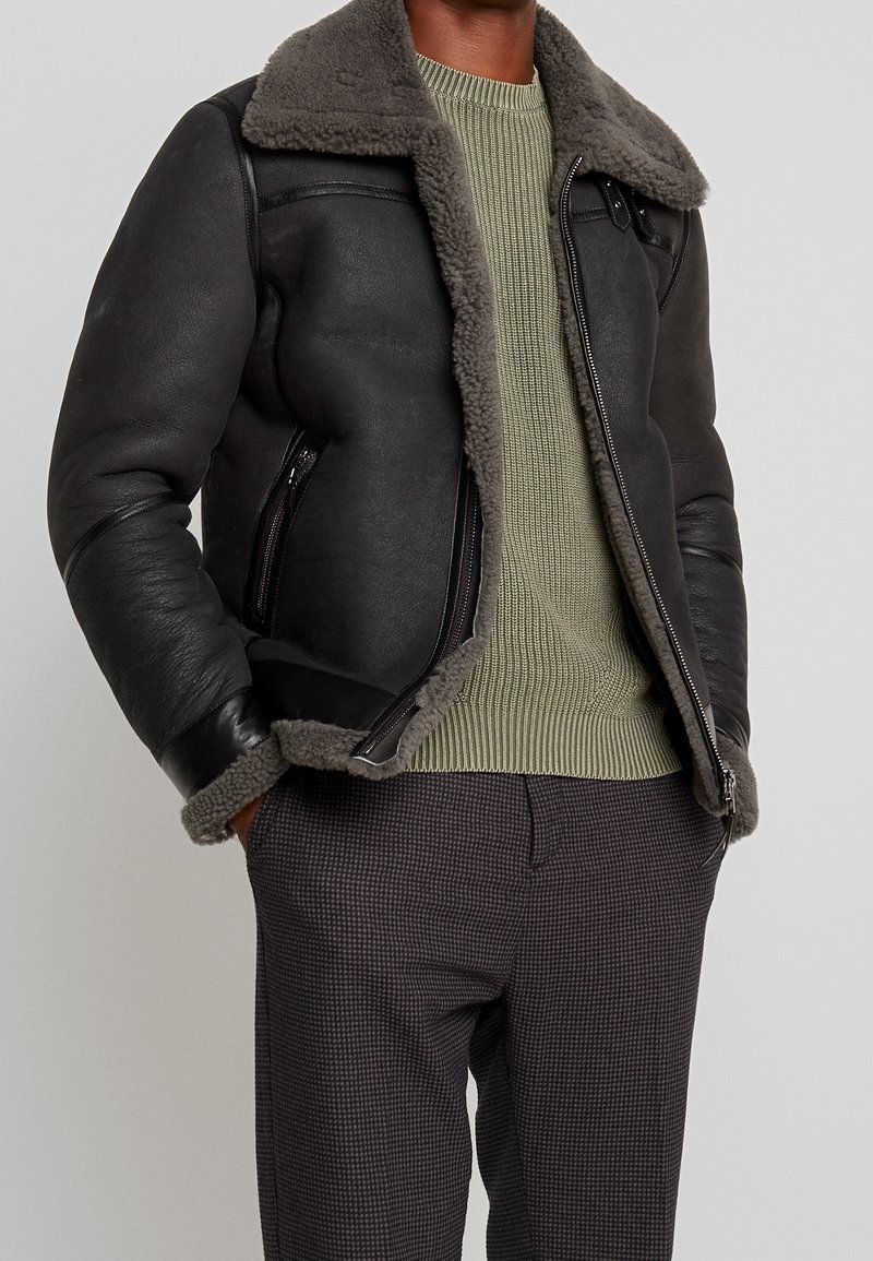 Giacca di pelle nera con colletto in shearling grigio, tasche con zip e design aderente. Indossata sopra un maglione verde chiaro e pantaloni a fantasia.