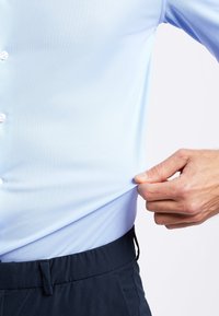 Lichtblauw, slim-fit overhemd met een gestructureerde stof, voorzien van witte knopen, gestoken in donkerblauwe broek, hand die aan de zoom van het shirt trekt.