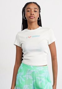 T-shirt blanc, coupé, avec "I ❤️ SURF" en texte bleu. Associé à un pantalon léger vert clair à motifs floraux et en tissu texturé.