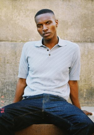Jeune homme aux cheveux courts portant un polo bleu clair texturé et un jean foncé, assis contre un mur en béton, regardant droit devant.