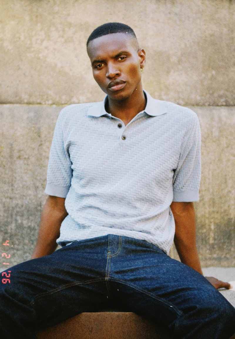 Jeune homme aux cheveux courts portant un polo bleu clair texturé et un jean foncé, assis contre un mur en béton, regardant droit devant.