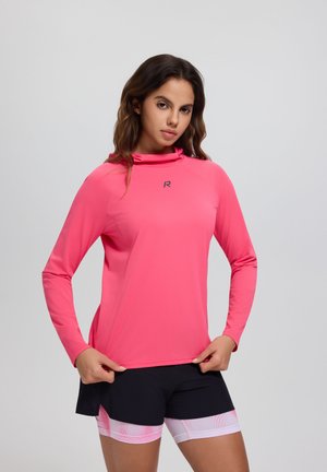 Frau trägt ein hellrosa langärmliges Sportoberteil und schwarze Shorts mit pink gemusterten Kompressionsleggings vor einfachem Hintergrund.