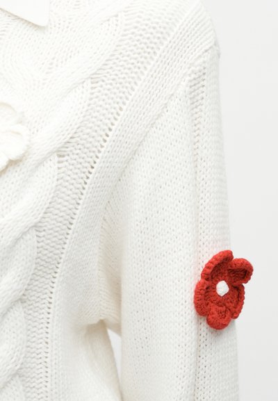 Hvit strikkegenser med mønstret tekstur, har en hekla rød blomst med hvitt sentrum på venstre erme.
