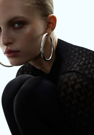 Close-up of a woman with slicked-back hair wearing large silver hoop earrings and a black textured top, looking intently ahead.