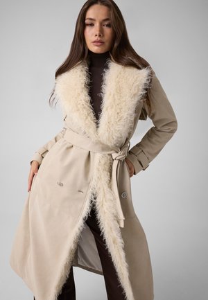 Femme portant un manteau beige ceinturé avec un grand col crème duveteux et un col roulé marron, debout les mains sur les hanches contre un fond gris uni.