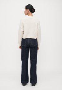 Kremfarget, kort strikkegenser med ribbede mansjetter. Mørkeblå vide jeans med synlige baklommer og kontrastsøm. Flate sko.