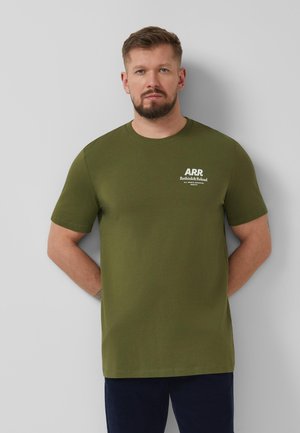 Brodaty mężczyzna z krótkimi włosami, ubrany w oliwkowozielony t-shirt z białym napisem „ARR Rethink & Reload”, stoi na tle jasnoszarej ściany.