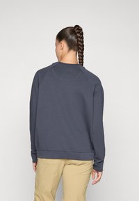 Mulher com um longo rabo de cavalo trançado, vestindo um moletom cinza-escuro e calças bege, de pé com as costas viradas para a câmara.