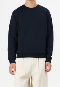 Mörkblå crewneck-tröja i mjukt tyg med långa ärmar, ribbad halsringning och avslappnad passform, kombinerad med ljusfärgade byxor.